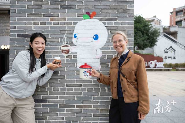 来自英国的游客(右)手持十五运特色杯子与喝咖啡的“喜洋洋”墙绘合影。